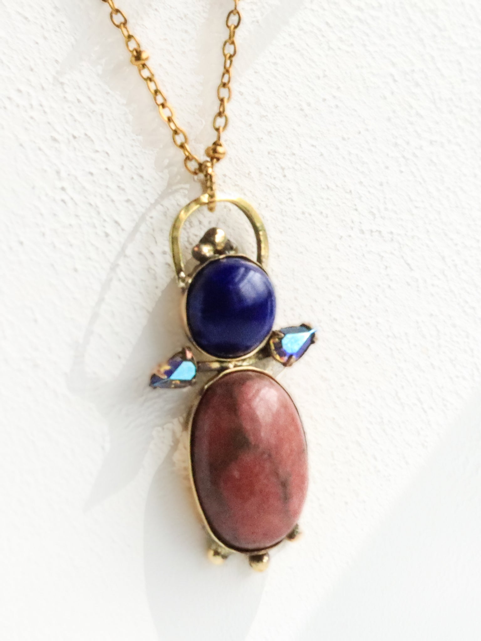 Close-up of handmade brass pendant featuring rhodonite cabochon, lapis lazuli stone and Swarovski comet crystal accents, bykjones artisan jewelry North Carolina