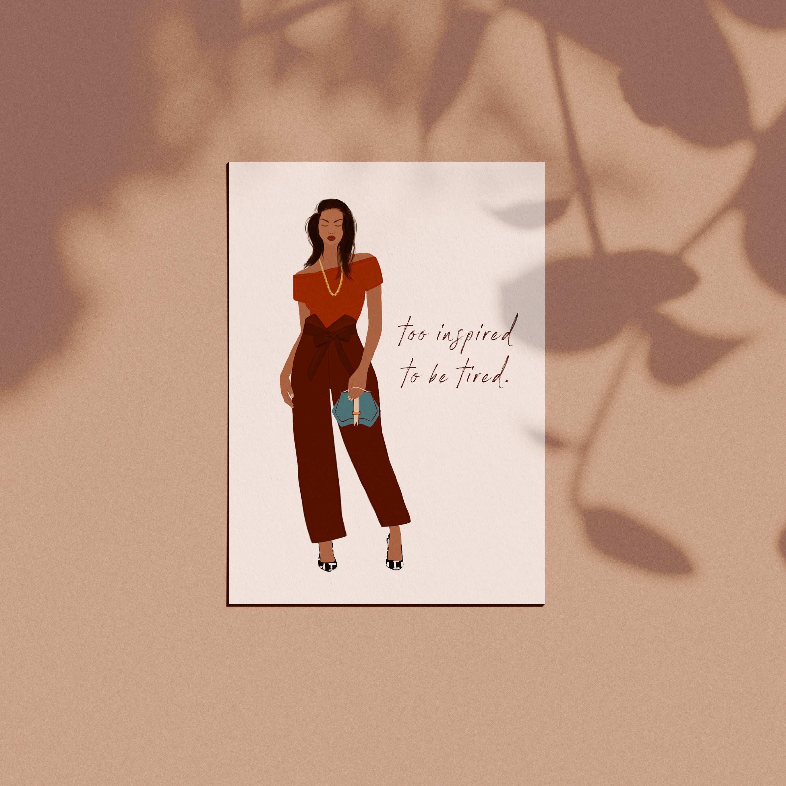 Close-up of a greeting card featuring a stylish woman in a rust top and brown pants, with the motivational quote "Too Inspired to Be Tired" against a minimalist design